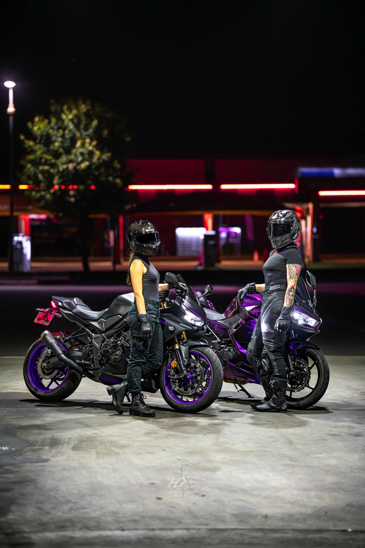 Deux femmes motardes posent l'une à côté de l'autres avec leurs motos à côté, de nuit, dans une station essence.