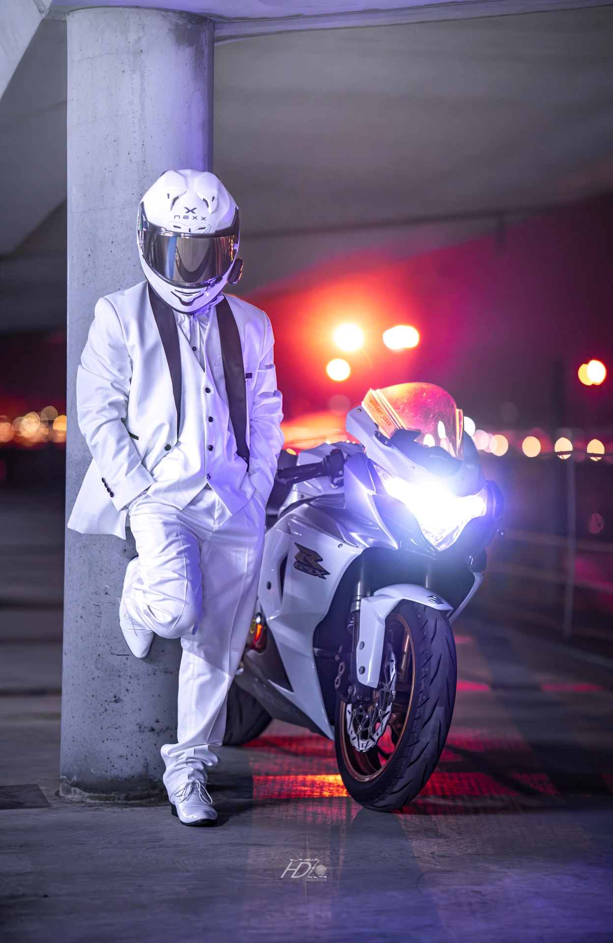 Un motard habillé tout en blanc pose sur un mur avec sa moto à côté.