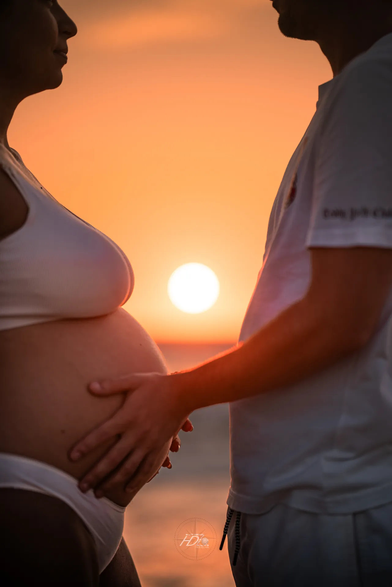 Un détail des mains sur le ventre de la femme enceinte lors d'une séance photo à la plage à côté de Arcachon avec le coucher de soleil en fond